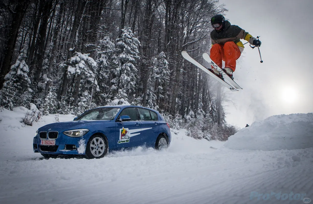 A blue BMW parked on a snow road as a skier jumps overhead through a forest.