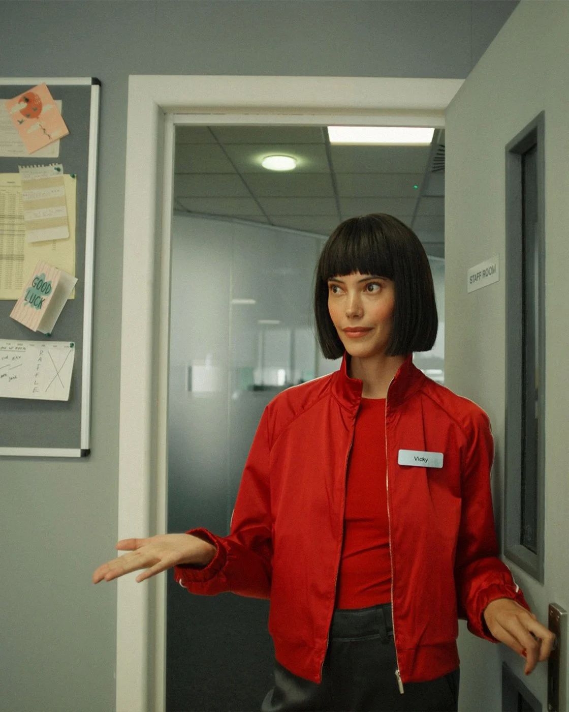 Film still: a woman in a red jacket with a Vicky name badge in an office doorway.